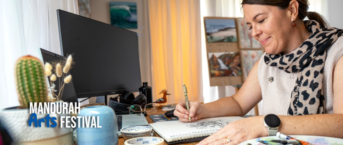 a woman sits at a desk cluttered with paints and paper, sketching in a notebook